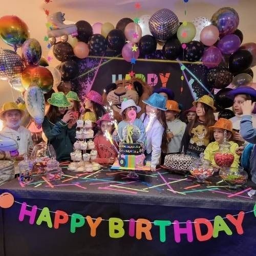 Children wearing colorful hats at a birthday party with balloons and a cake on the table.