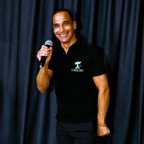 Person wearing a black shirt with a logo, holding a microphone in front of a dark curtain.