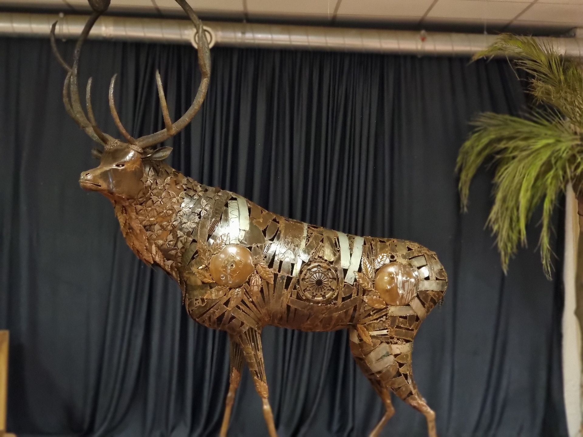 Sculpture of a stag made from various metal pieces, displayed against a dark curtain backdrop.