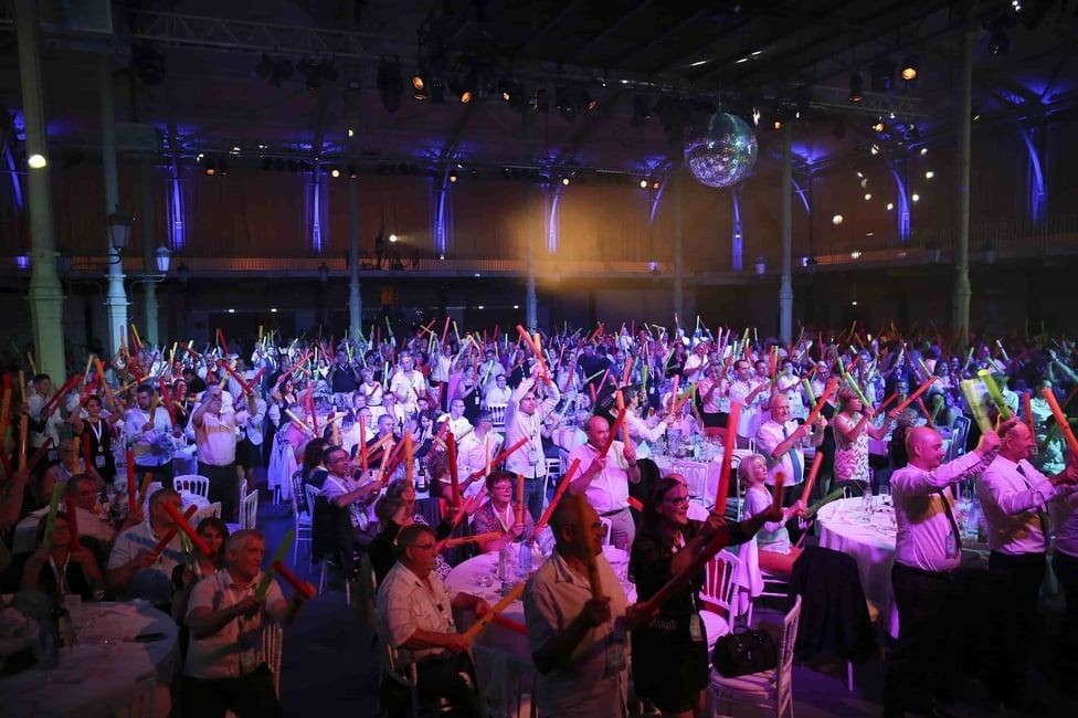 Large crowd at event holding colorful glow sticks, with purple lighting and a disco ball overhead.